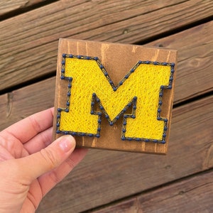 May include: A wooden plaque with a yellow string art letter M. The letter is outlined in navy blue.
