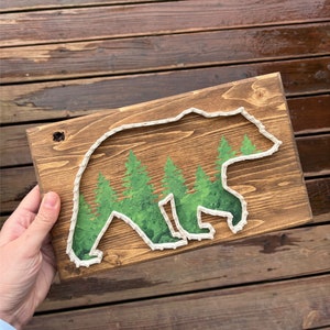 May include: A wooden sign with a white string art design of a bear walking through a forest. The bear is outlined in white string and the forest is depicted with green paint.