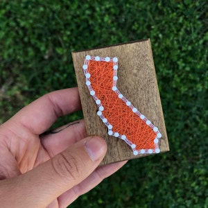 May include: A wooden plaque featuring a string art design of the state of California. The outline is made with white nails, and the interior is filled with orange string. The wood has a natural, stained finish.