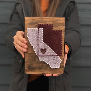 May include: A wooden sign with a string art design of the state of California. The state is outlined in white string and filled in with maroon string. A small heart is in the center of the state.