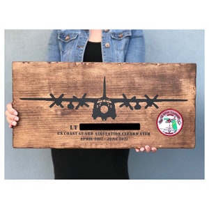 May include: A wooden sign with a black silhouette of a C-130 Hercules aircraft. The sign reads "LT [blank] US Coast Guard Air Station Clearwater April 2017 - June 2021". A red, green, and white patch with the words "US Coast Guard Air Station Clearwater" is in the upper right corner.