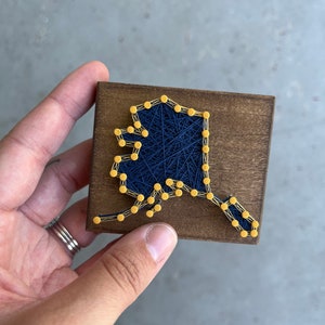 May include: A wooden plaque with a string art design of the state of Alaska. The outline of the state is created with yellow tacks and blue string.