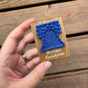 May include: A small wooden plaque with a blue string art design of the Liberty Bell. The plaque has the text "Heritage Foundation" printed on it.