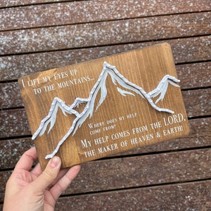 May include: Wooden sign with a white string art mountain design and the text "I lift my eyes up to the mountains... Where does my help come from? My help comes from the Lord, the maker of heaven & earth!"
