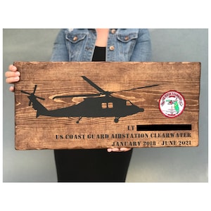 May include: A wooden sign with a black silhouette of a helicopter, the text "US Coast Guard Air Station Clearwater", "January 2018 - June 2021", and "LT [blank space]" and a red, white, and blue patch with a helicopter and the text "US Coast Guard" and "Air Station Clearwater".