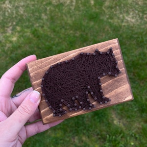 May include: A brown wooden plaque with a brown string art bear design. The bear is outlined with small nails and the string is wrapped around the nails to create the bear shape.