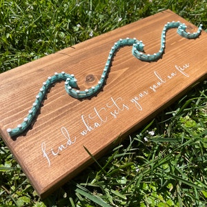 May include: A wooden sign with a teal string art design of waves and the text "find what sets your soul on fire".
