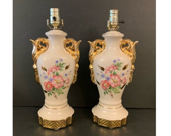 Może przedstawiać: Para białych ceramicznych lamp stołowych z złotymi akcentami i wzorami kwiatowymi. Lampy mają klasyczny kształt urny ze złotą podstawą i wzorem kwiatowym z różowymi i niebieskimi kwiatami.