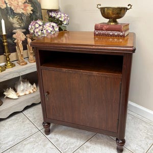May include: A dark brown wooden nightstand with a single door and a small open shelf. The nightstand has a glossy finish and sits on four rounded legs. A brass bowl and books are on top.