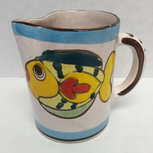 May include: A white ceramic pitcher with a blue and brown rim and a yellow fish with red and green details painted on the side. The pitcher has a brown handle.