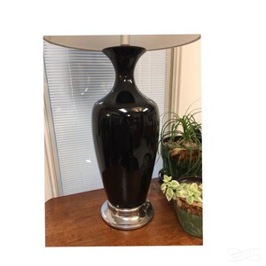 May include: A black ceramic table lamp with a silver base and a white lampshade. The lamp is sitting on a wooden table.