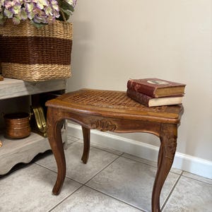 May include: A small, antique wooden side table with a woven top and curved legs. The table is stained a warm brown color and features carved details. Two vintage books rest on the table. The table is a decorative furniture piece.