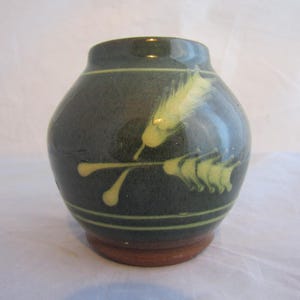 May include: A small, dark green ceramic vase with a white and yellow floral design. The vase has a rounded body and a slightly flared lip.