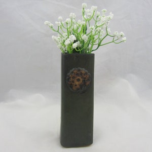 May include: A tall, rectangular dark gray ceramic vase featuring a circular, textured brown medallion on the front. The vase is filled with a cluster of small, delicate white flowers on green stems, creating a contrast against the dark vessel.