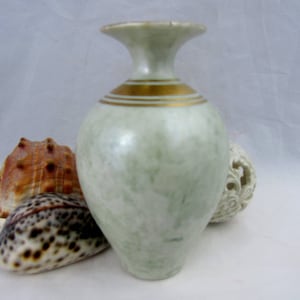 May include: A small, light green ceramic vase with a gold band around the top. The vase has a pearlescent finish and is sitting on a white surface.