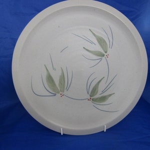 May include: A round, off-white ceramic plate with a speckled texture. The surface features a painted design of three clusters of green leaves, thin blue lines representing stems or needles, and small red dots resembling berries arranged in a circular pattern.