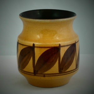 Könnte beinhalten: Eine Keramikvase mit bauchigem Körper und ausgestelltem Rand. Die Außenseite ist in einem warmen Gelb-Beige glasiert und mit einem horizontalen Band aus dunkelbraunen Blattmotiven verziert. Das Innere der Vase ist dunkelbraun glasiert.