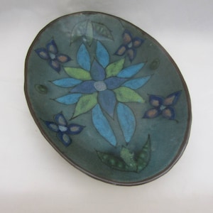 May include: A teal ceramic oval dish with a blue and green floral design. The dish has a brown rim and is decorated with a large blue flower in the centre and smaller blue flowers around the edge.