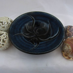 May include: A circular dark blue ceramic bowl with a glossy finish and a raised, swirling black floral-like relief pattern in the center. The bowl is accompanied by two off-white spherical decorative objects and two natural sea shells.