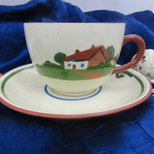 Könnte beinhalten: Eine weiße Teetasse und Untertasse aus Keramik mit handbemaltem Motiv eines roten Backsteinhauses mit grünen Bäumen und einem braunen Weg. Die Tasse hat einen braunen Henkel und die Untertasse einen grünen und blauen Rand.