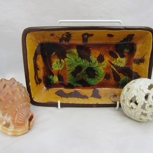 May include: A rectangular ceramic tray with a glossy mustard yellow glaze, dark brown rim, and abstract dark brown and green splattered patterns. Beside it sits a tan, ridged seashell and a spherical, off-white carved decorative object.