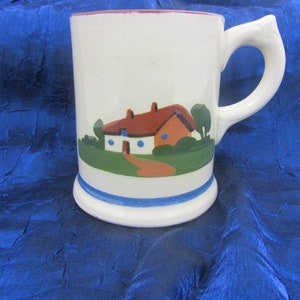 May include: White ceramic mug with a whimsical illustration of a house with a red roof, green grass, and a path. The mug has a unique handle design and a blue stripe near the base. The rim is trimmed in red.