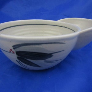 May include: A speckled off-white ceramic bowl with a side handle and a rounded shape. The exterior features a hand-painted design of dark gray leaves and three small red dots. A thin dark gray line circles the interior rim of the bowl.