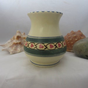 May include: A cream-colored ceramic vase with a flared rim and a rounded body. The vase features a horizontal band of dark green with a repeating brown vine pattern containing small red three-petal flower motifs and blue trim accents.