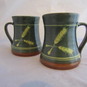 May include: Two green ceramic mugs with yellow wheat designs. The mugs have a brown rim and base.