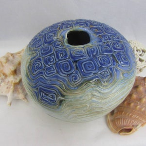 May include: A blue and white ceramic vase with a swirling pattern. The vase has a round shape and a small opening at the top. It is sitting on a white surface with seashells.