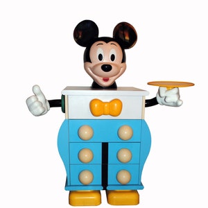 Peut inclure: Une commode bleue et blanche avec une tête de Mickey Mouse sur le dessus. La commode a quatre tiroirs avec des boutons blancs et un nœud papillon jaune sur le tiroir du haut. La tête de Mickey Mouse sourit et a des oreilles noires et un nez noir.