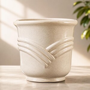 May include: A beige ceramic planter with a speckled finish. The planter features a decorative, raised design around the middle. The top rim curves outward, and the base is slightly flared. A stylish addition to any home decor.