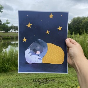 May include: A print featuring a sleeping koala under a yellow blanket against a dark blue starry night sky. The koala is nestled on a purple pillow. The artist's signature, "kathy osborne studio," is at the bottom.