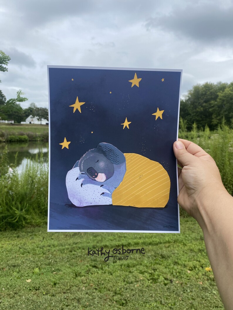 May include: A print featuring a sleeping koala under a yellow blanket against a dark blue starry night sky. The koala is nestled on a purple pillow. The artist's signature, "kathy osborne studio," is at the bottom.