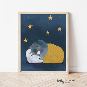 May include: A framed art print featuring a sleeping koala under a yellow blanket against a dark blue starry night sky. The koala is gray with a pink nose, resting on a white pillow. The artist's name, "kathy osborne studio", is in the bottom right corner.