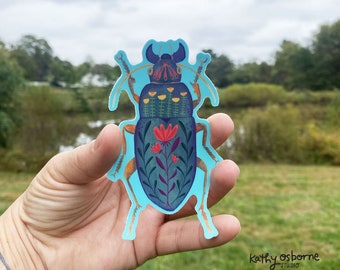 May include: A blue and green beetle sticker with floral details. The beetle has red flowers and yellow flowers on its back. The sticker is on a light blue background.