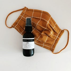 May include: A black glass bottle with a white label that reads "GOOD FACE MIST" and "relief" in a smaller font. The bottle is sitting on top of a brown and white checkered fabric face mask.