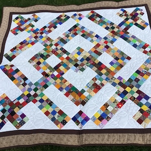 May include: A white quilt with a brown border. The quilt is made up of small squares of fabric in various colors and patterns. The quilting design features a repeating pattern of leaves and flowers.