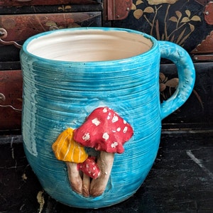 May include: A blue ceramic mug with a handle and a three-dimensional design of red and yellow mushrooms.