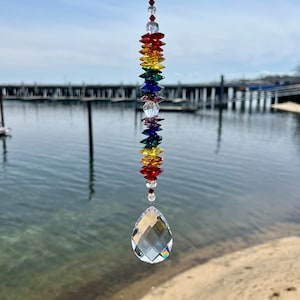 May include: A rainbow crystal suncatcher with a teardrop-shaped crystal at the bottom. The suncatcher is hanging from a white string and is in front of a blurry background of a body of water and a wooden pier.