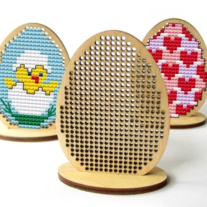 Könnte beinhalten: Drei hölzerne, eiförmige Stickrahmen, zwei davon mit fertigen Kreuzstichmustern. Ein Rahmen zeigt ein gelbes Küken in einem weißen Ei mit blauen und grünen Akzenten. Der zweite Rahmen hat ein rosa-weiß gestreiftes Design. Der dritte Rahmen ist leer.