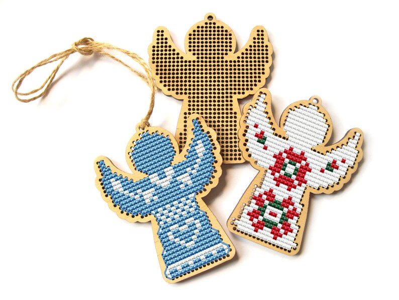May include: Three wooden angel ornaments with cross-stitch designs. One angel features a blue and white pattern, another has a red and green design, and the third is unfinished. Each has a jute string for hanging.