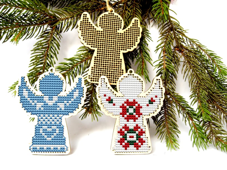 May include: Three wooden angel ornaments with cross-stitch designs, displayed on a bed of evergreen branches. One angel is plain wood, while the others feature blue and red patterns. Each has a small hole for hanging.