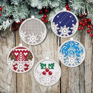 Op de afbeelding: Vijf ronde kerstversieringen met witte sneeuwvlokuitsparingen. De ornamenten hebben verschillende ontwerpen, waaronder een raster, een blauw kruissteekpatroon, een rood-groen patroon, hulstbessen en een blauw-wit kruissteekpatroon. De achtergrond is een houten oppervlak.