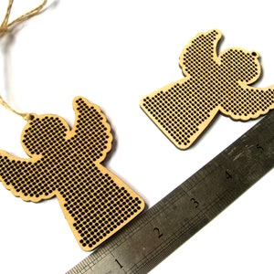May include: Two wooden angel ornaments with a perforated design. One angel is attached to a twine string. A metal ruler is placed diagonally across the image. The angels are made of light-colored wood and have a detailed cutout pattern.