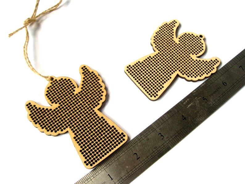 May include: Two wooden angel ornaments with a perforated design. One angel is attached to a twine string. A metal ruler is placed diagonally across the image. The angels are made of light-colored wood and have a detailed cutout pattern.