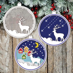 May include: Three round Christmas ornaments with white deer silhouettes. One ornament has a white mesh background, another has a blue background with stars, and the third has a blue background with a village scene. The ornaments are hanging on a wooden background.