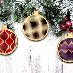 Op de afbeelding: Drie houten kerstversieringsvormen met kruissteekpatronen. De ornamenten zijn rood, paars en zwart met gouden accenten. De ornamenten bevinden zich op een witte houten achtergrond.