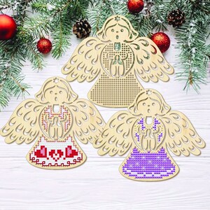 Peut inclure: Trois ornements en bois en forme d'ange, avec des ailes et des visages d&eacute;coup&eacute;s. Chaque ange pr&eacute;sente un motif de point de croix diff&eacute;rent en rouge, blanc et violet. Les anges sont pr&eacute;sent&eacute;s sur une surface en bois blanc avec des d&eacute;corations de No&euml;l.