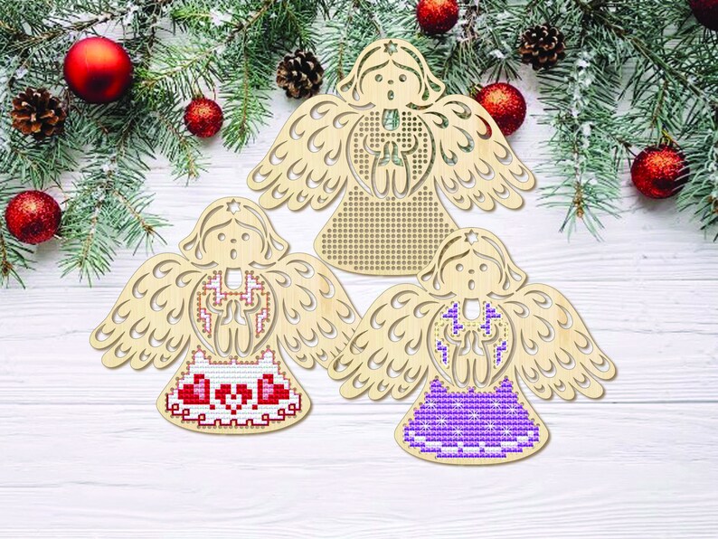 Peut inclure: Trois ornements en bois en forme d'ange, avec des ailes et des visages d&eacute;coup&eacute;s. Chaque ange pr&eacute;sente un motif de point de croix diff&eacute;rent en rouge, blanc et violet. Les anges sont pr&eacute;sent&eacute;s sur une surface en bois blanc avec des d&eacute;corations de No&euml;l.